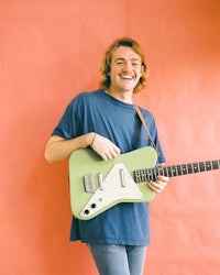 a young man holding a green electric guitar in front of a pink wall