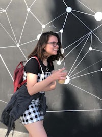 a girl wearing a plaid dress and carrying a cup of coffee in front of a black wall